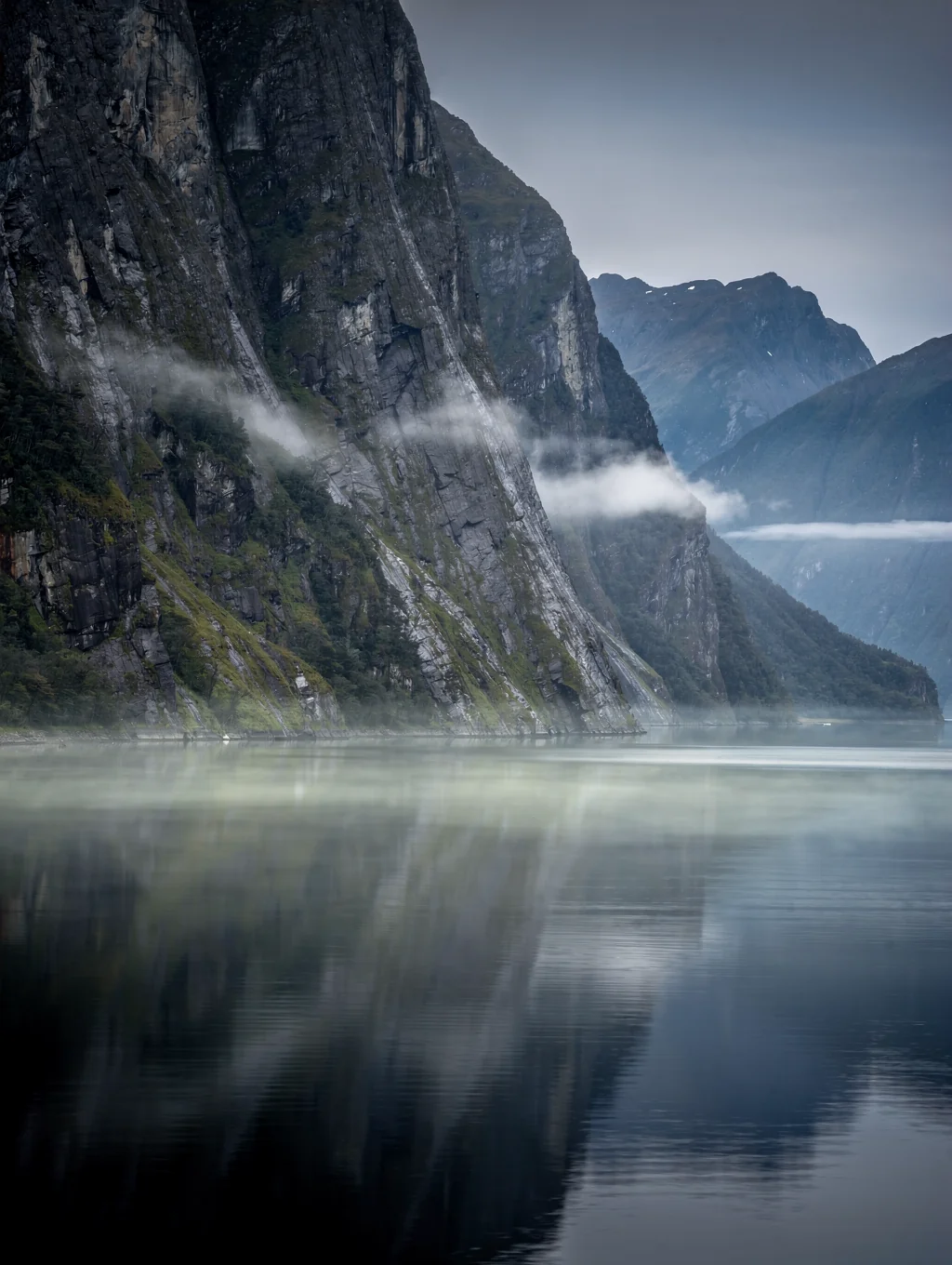 Cascading rainforest cliff waterfalls into dark fjord waters in New Zealand